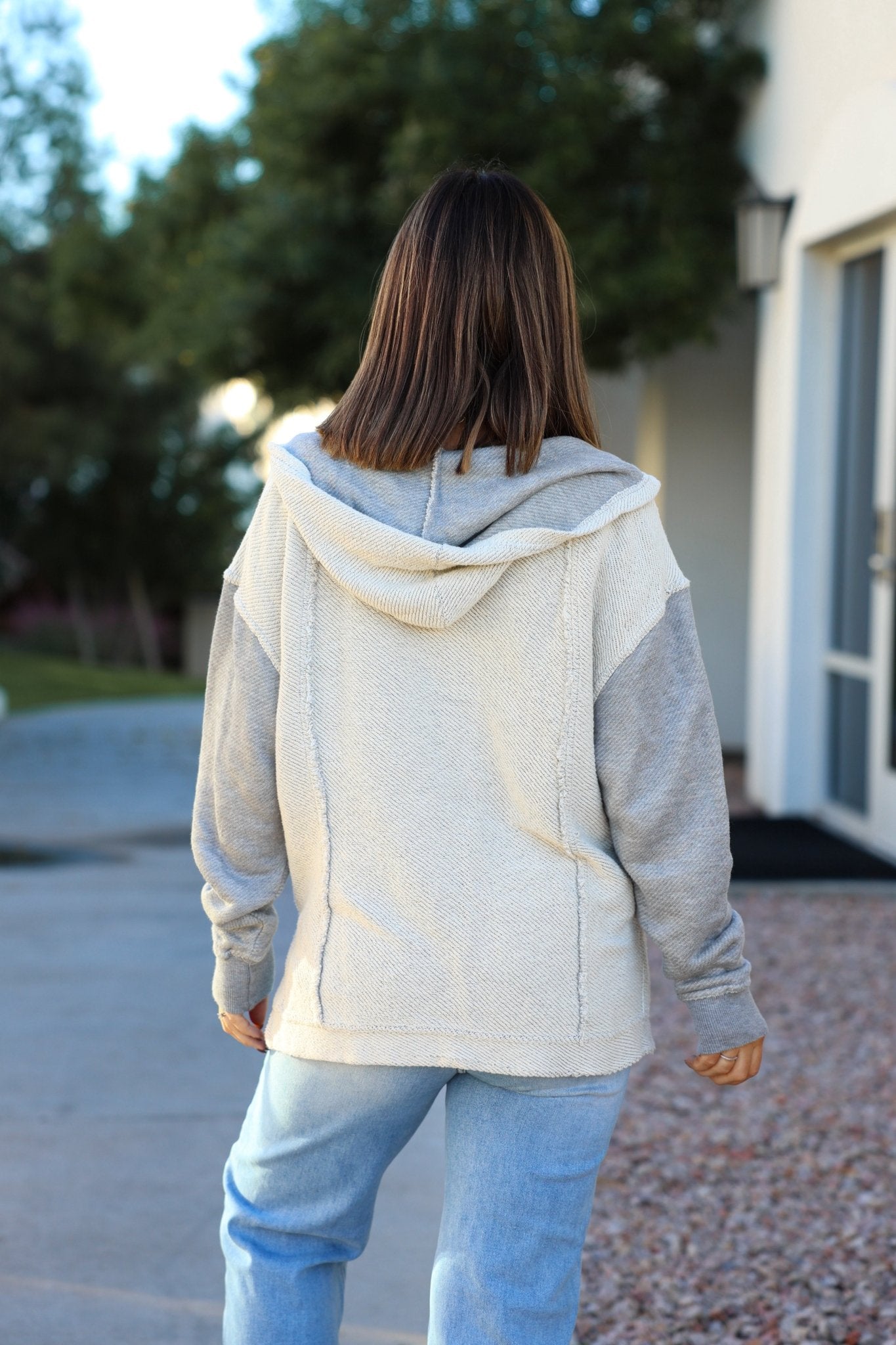 A person in a Heather Grey Slouchy Hooded Sweatshirt and jeans, with straight brown hair, stands outdoors facing away.