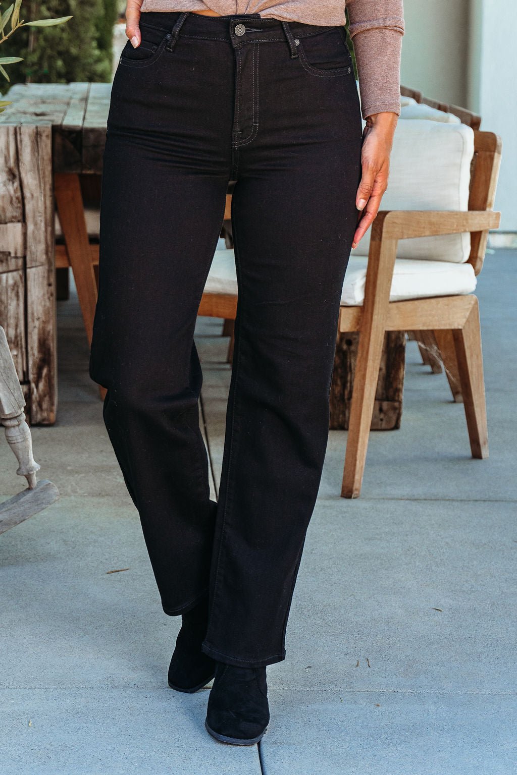 A person in Hidden Black High Rise Straight Leg Jeans and black boots stands outdoors near wooden furniture.