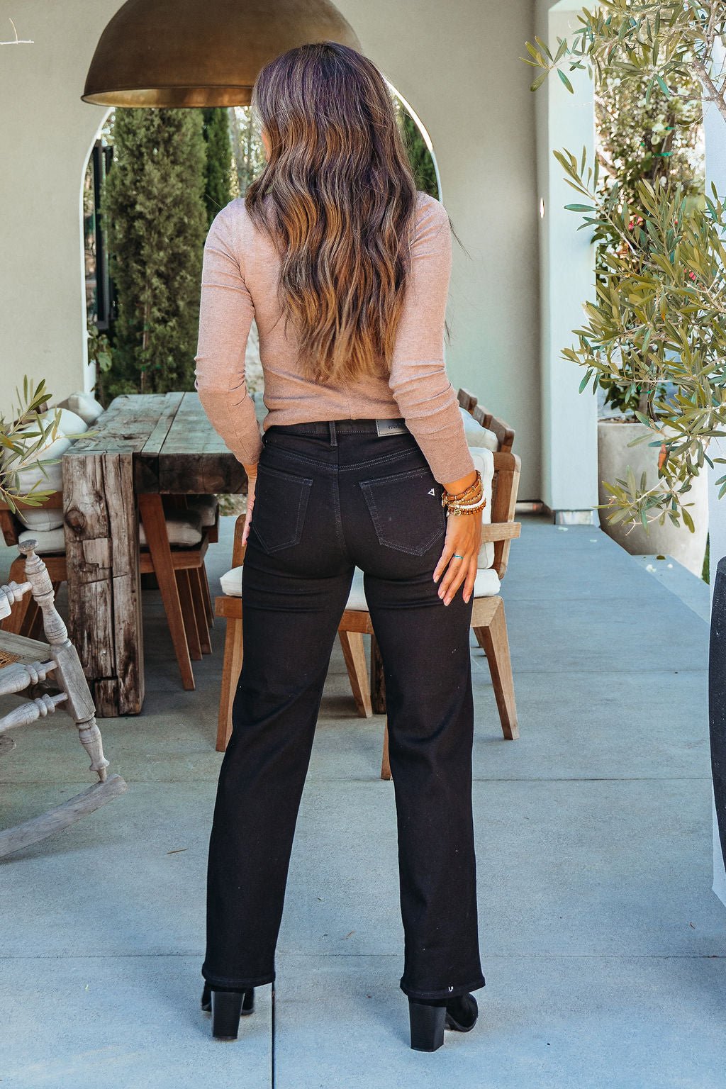 A woman with long wavy hair in a tan top and Hidden Black High Rise Straight Leg Jeans stands with her back to the camera outdoors.