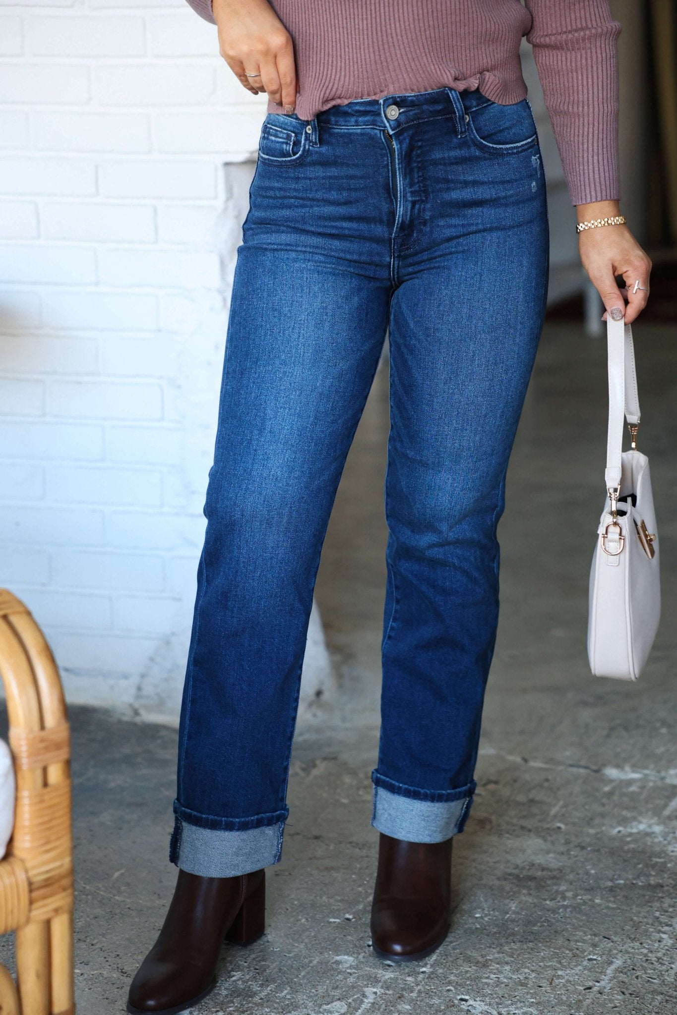 Person wearing Hidden Dark Wash High Rise Cuffed Jeans, a mauve sweater, dark boots, and holding a white handbag by a white brick wall.