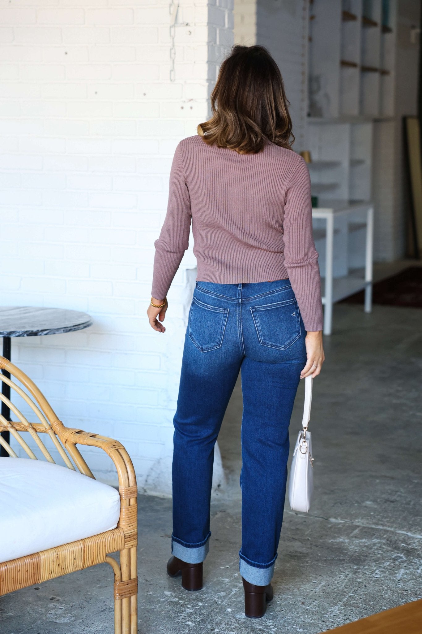 A woman in Hidden Dark Wash High Rise Cuffed Jeans stands indoors, facing away, holding a light-colored handbag.
