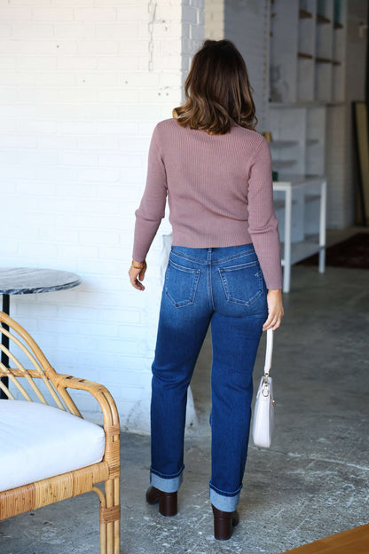 A woman in Hidden Dark Wash High Rise Cuffed Jeans stands indoors, facing away, holding a light-colored handbag.