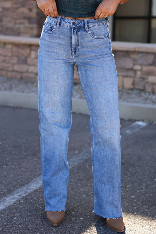 Model in Hidden Light Wash Basic Dad Jeans stands on pavement, wearing brown pointed shoes with a stone wall as backdrop.