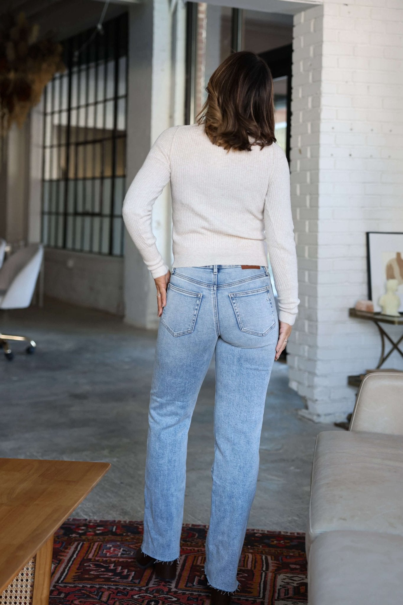 A woman wears Hidden Light Wash Straight Leg Jeans and a white sweater, standing indoors in a modern living room with white brick walls.