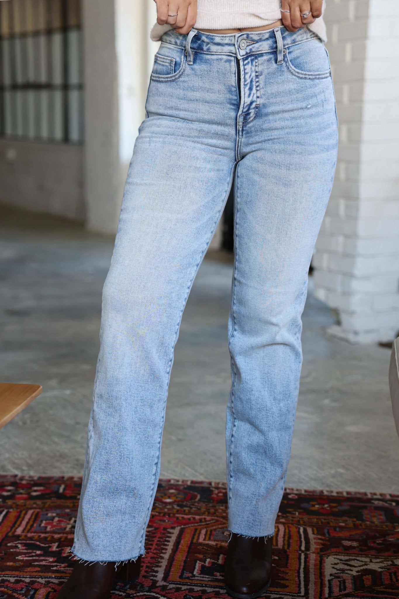 Person wearing Hidden Light Wash Straight Leg Jeans with black boots, standing indoors on a rug by a white brick wall.