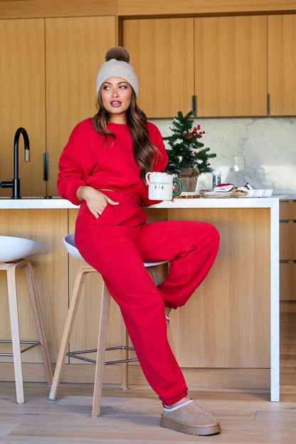 Woman in Holiday Red Lounge Jogger Sweatpants - FINAL SALE sits on a kitchen stool, holding a mug by a decorated Christmas tree.