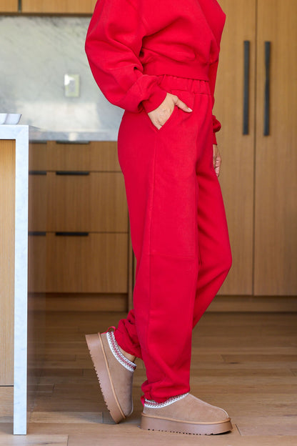 A person wears Holiday Red Lounge Jogger Sweatpants - FINAL SALE and tan slippers in a modern kitchen with wood cabinets and light floors.