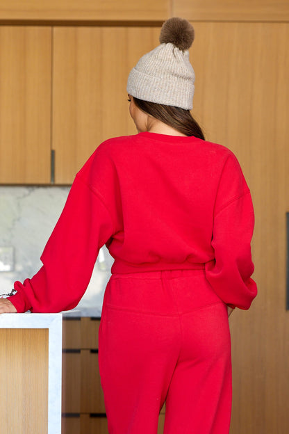 Wearing the Holiday Red Lounge Sweatshirt, a person in pants and a beige knit pom-pom hat stands with their back to the camera.