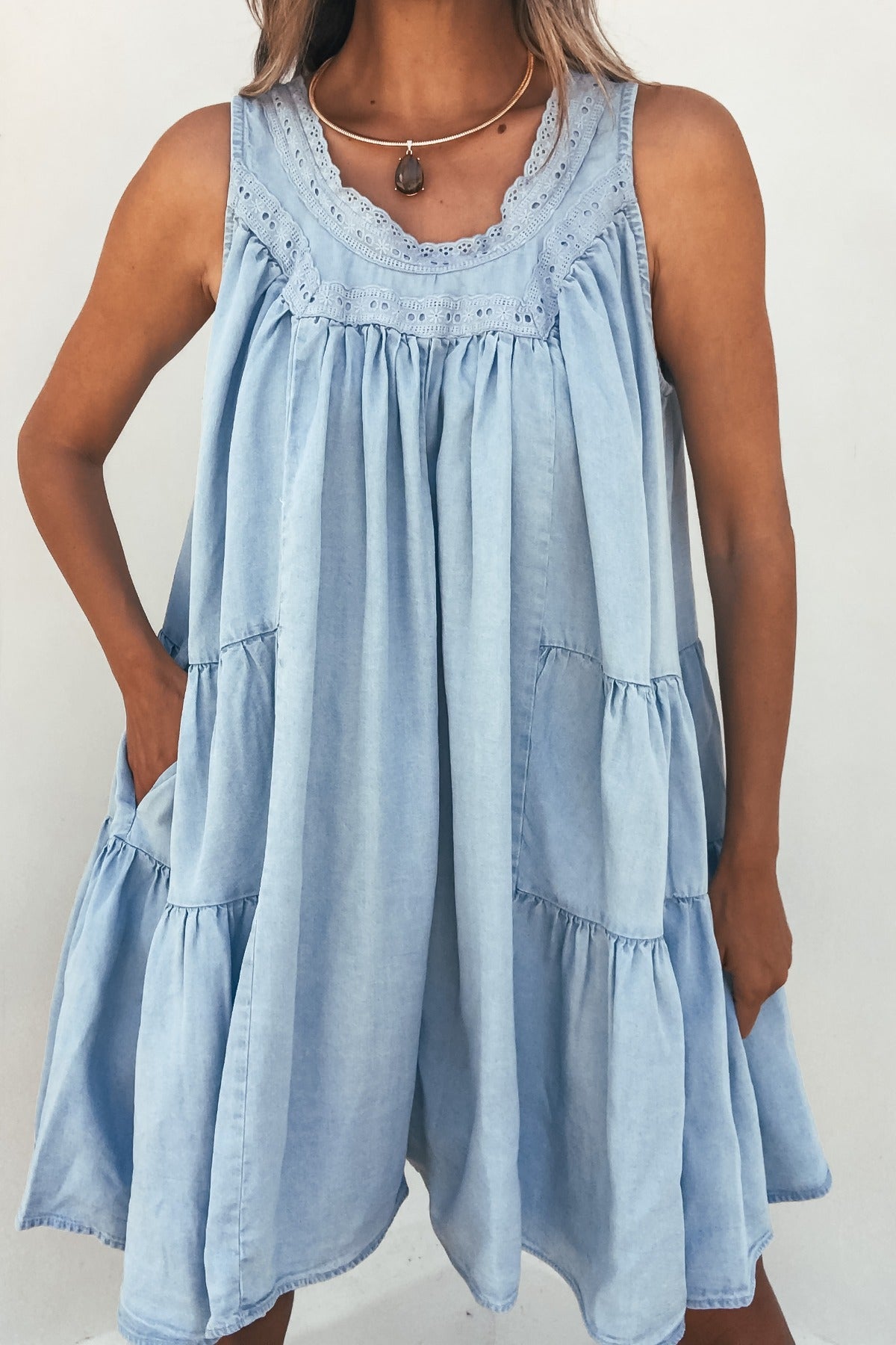 Woman wearing the Hollis Chambray Tiered Romper, a sleeveless light blue romper with lace trim, pockets, and relaxed fit.
