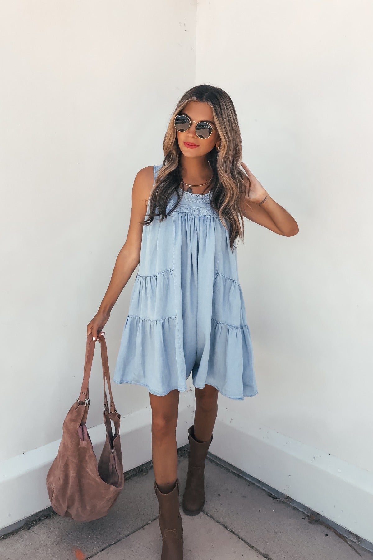 Woman in a Hollis Chambray Tiered Romper, brown boots, sunglasses, and a brown bag, standing casually by a white wall.