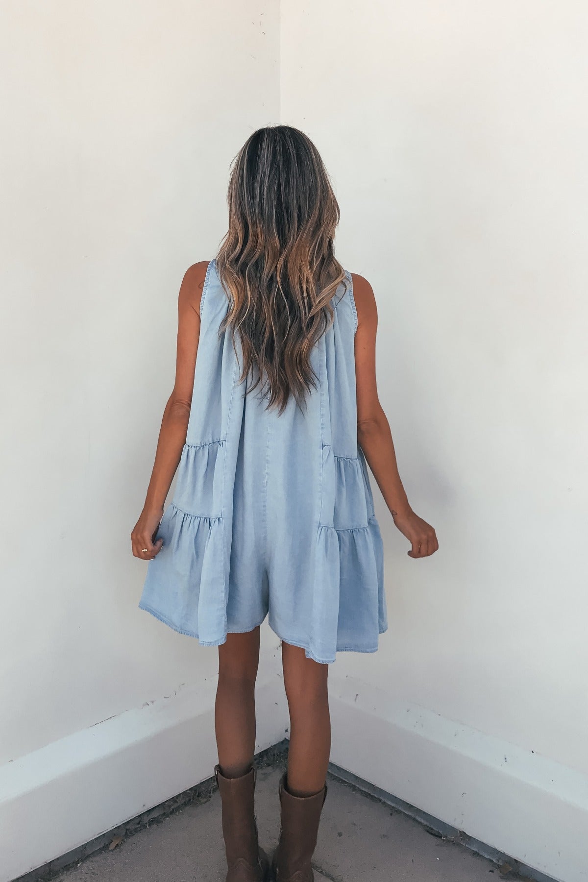 A woman with long wavy hair wears the Hollis Chambray Tiered Romper and brown boots, facing a white corner wall.