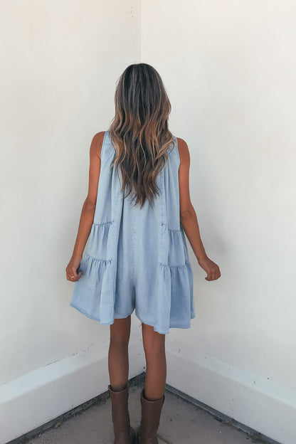 A woman with long wavy hair wears the Hollis Chambray Tiered Romper and brown boots, facing a white corner wall.