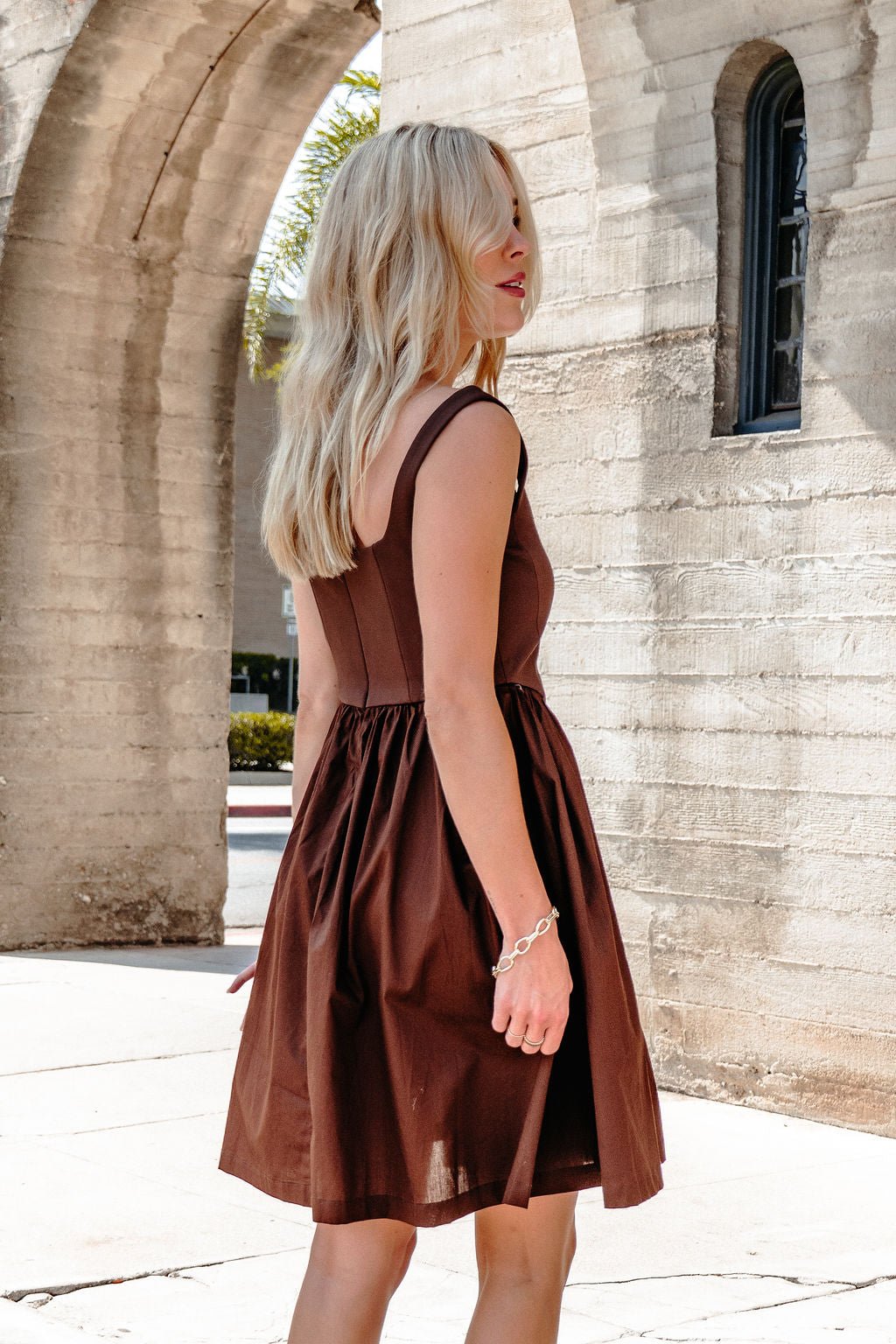 A woman with long blonde hair models the Hunter & Brown Corset Stretch Mini Dress near arched concrete walls, looking over her shoulder.