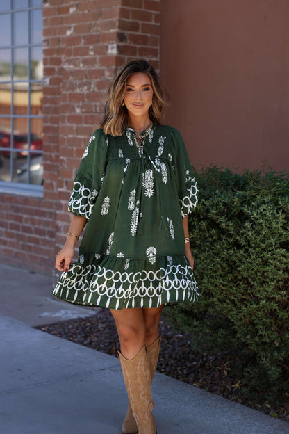 A woman in a Hunter Green Border Print Tiered Mini Dress smiles at the camera, standing outside by a brick wall and bushes.