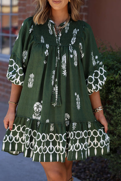 Woman in a Hunter Green Border Print Tiered Mini Dress stands by a brick wall and green bush.