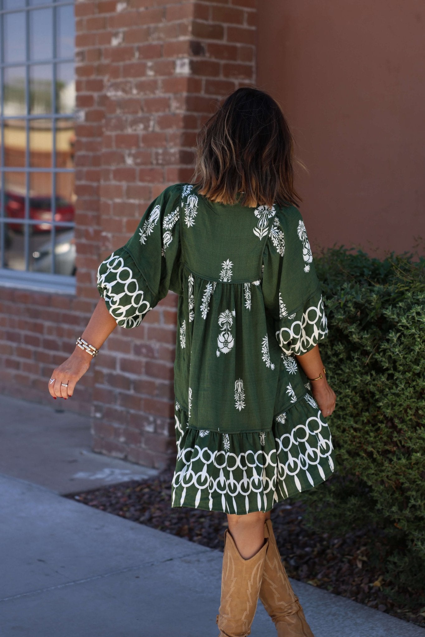 A woman in a Hunter Green Border Print Tiered Mini Dress and tan boots walks outside near a brick building, facing away.