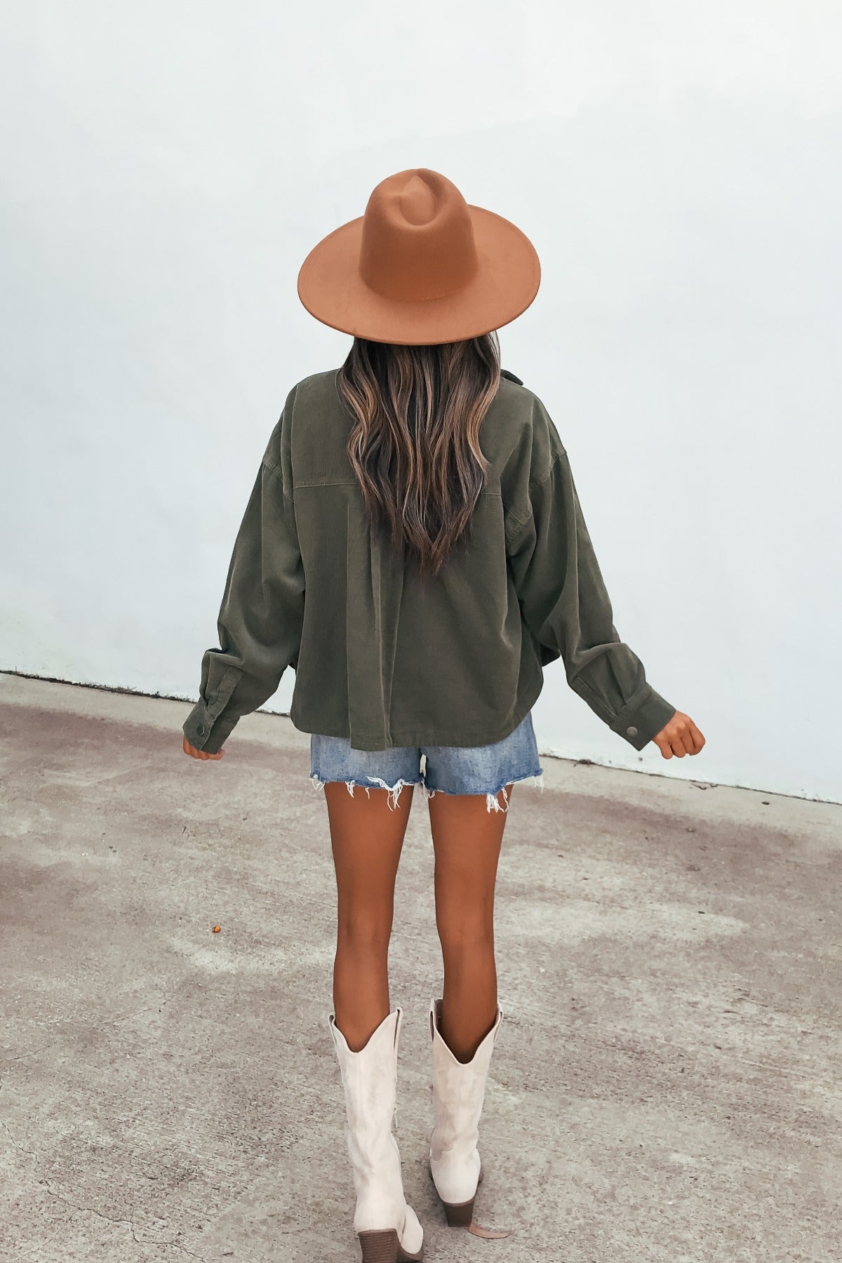 A woman in a brown hat, Hunter Green Corduroy Shacket, denim shorts, and white boots stands facing away on concrete.