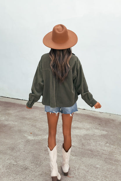 A woman in a brown hat, Hunter Green Corduroy Shacket, denim shorts, and white boots stands facing away on concrete.