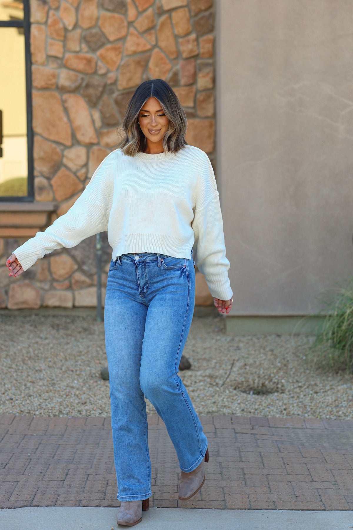 Woman with shoulder-length hair wears the HYFVE cream drop shoulder sweater, blue jeans, and boots, walking outside by a stone wall.