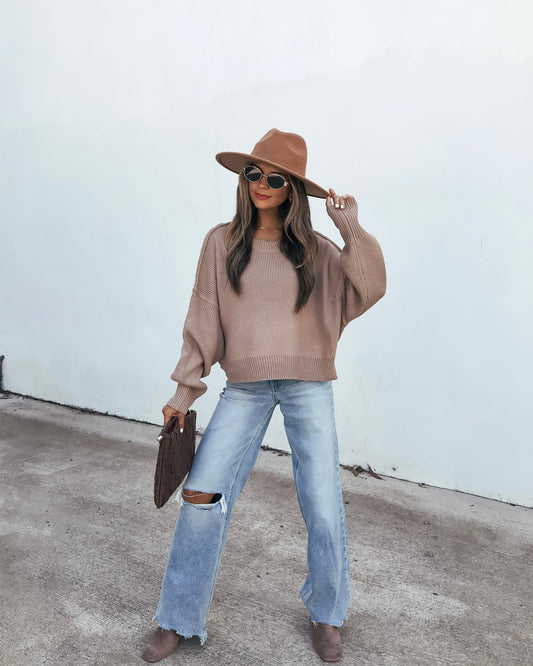 A person wearing a Ribbed Drop Shoulder Sweater - Taupe, distressed light-wash jeans, a wide-brim hat, and sunglasses while holding a clutch.
