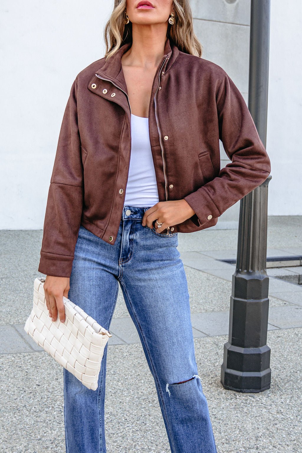 A woman wears a HYFVE HF26E373 CHESTNUT FAUX SUEDE ZIP UP JACKET over a white top and blue jeans, carrying a white woven clutch.
