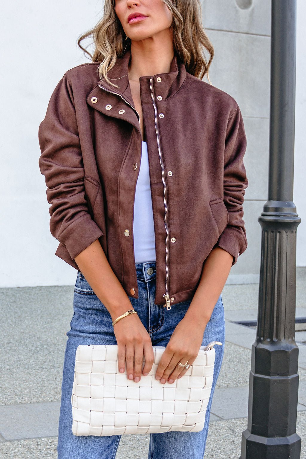 A woman wearing a HYFVE HF26E373 CHESTNUT FAUX SUEDE ZIP UP JACKET, white top, and blue jeans, holding a white woven clutch bag.