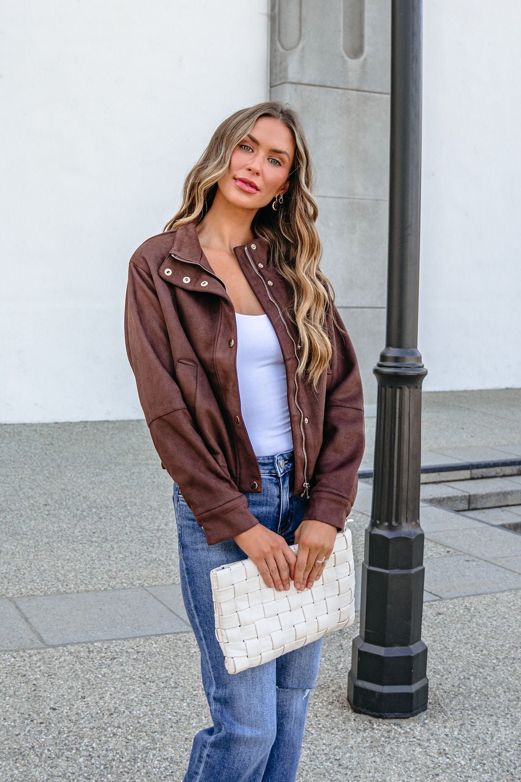 A woman wearing a HYFVE HF26E373 CHESTNUT FAUX SUEDE ZIP UP JACKET, white top, and blue jeans holds a white woven clutch.