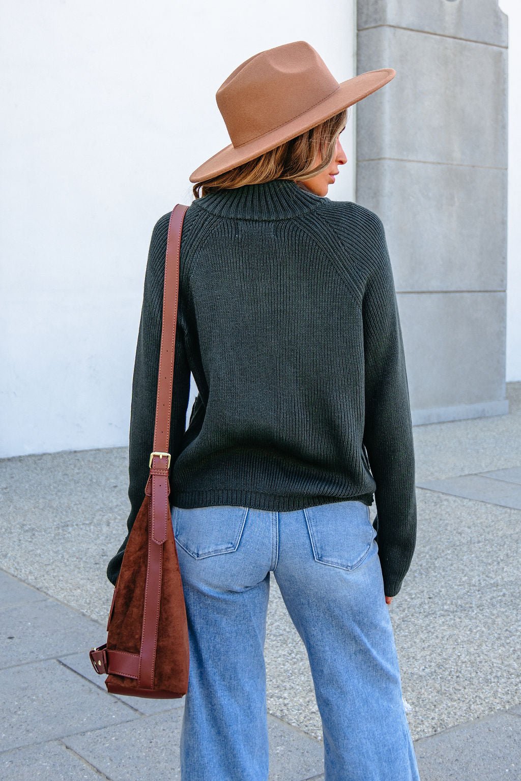 Woman in a brown hat, forest green zip up sweater, and jeans stands outdoors with her back to the camera, holding a shoulder bag.
