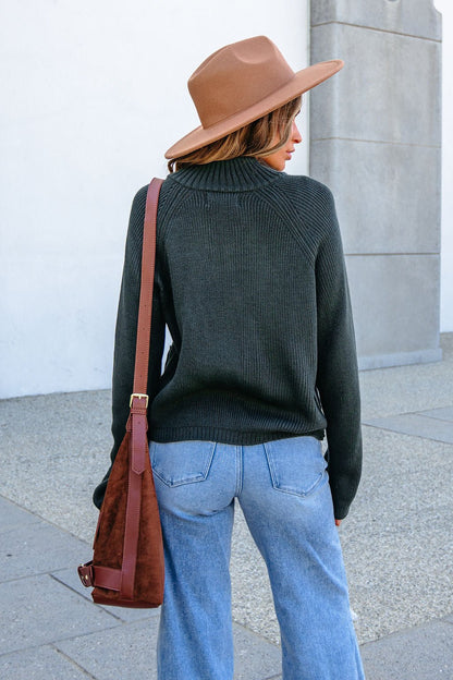 Woman in a brown hat, forest green zip up sweater, and jeans stands outdoors with her back to the camera, holding a shoulder bag.
