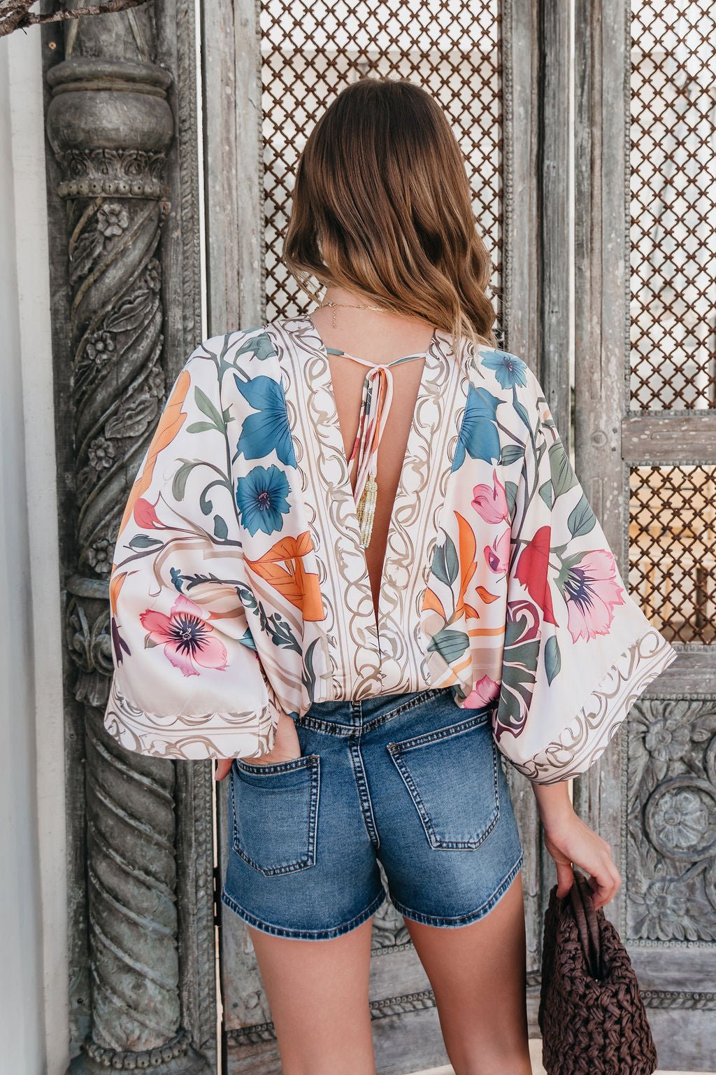 Woman with wavy hair wears the Island Muse Multi Floral Bodysuit and denim shorts, holding a brown bag by ornate wooden doors.