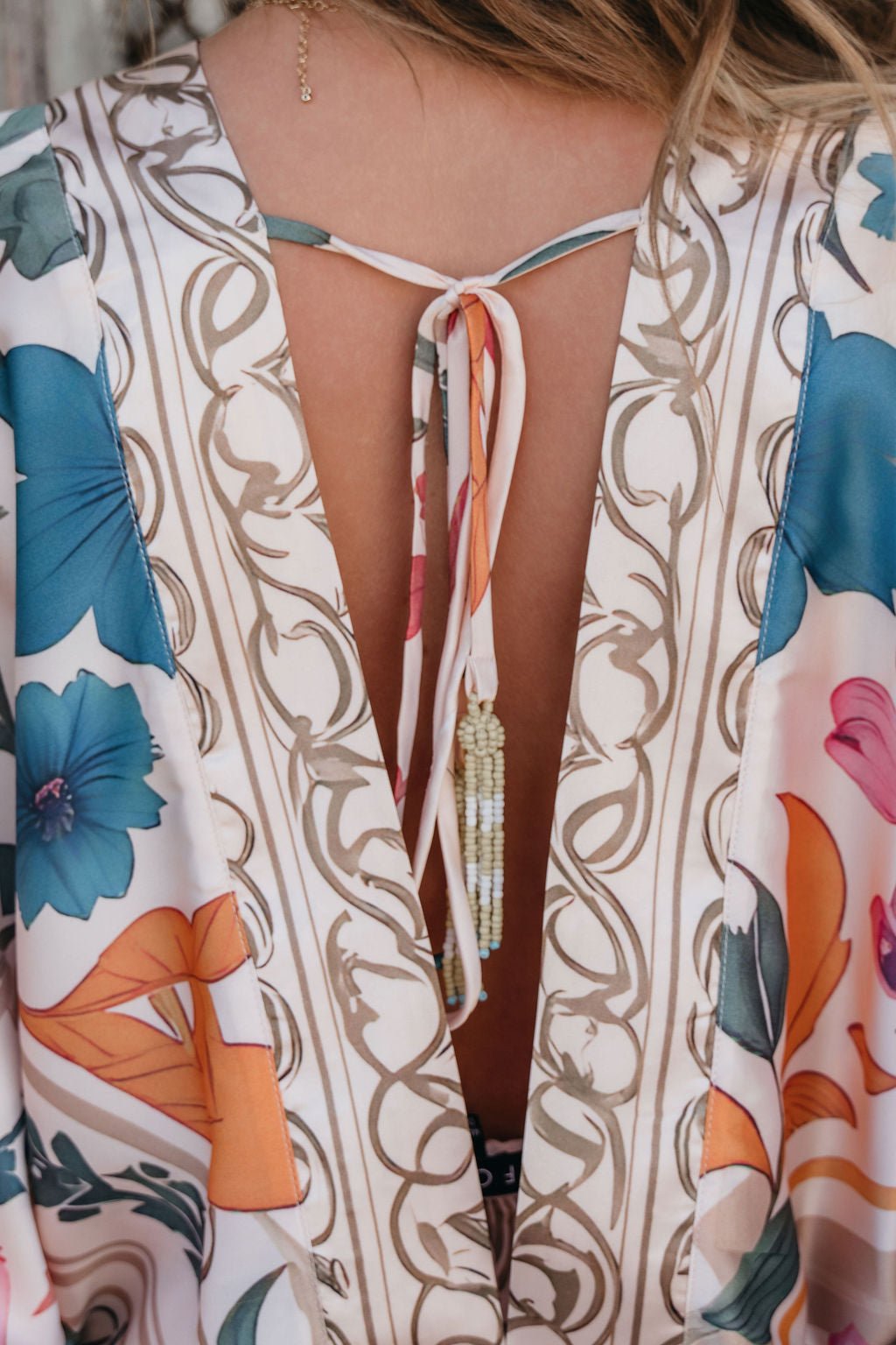 A close-up of a woman's back in the Island Muse Multi Floral Bodysuit with an open back, tie, beaded neckline, and kimono sleeves.