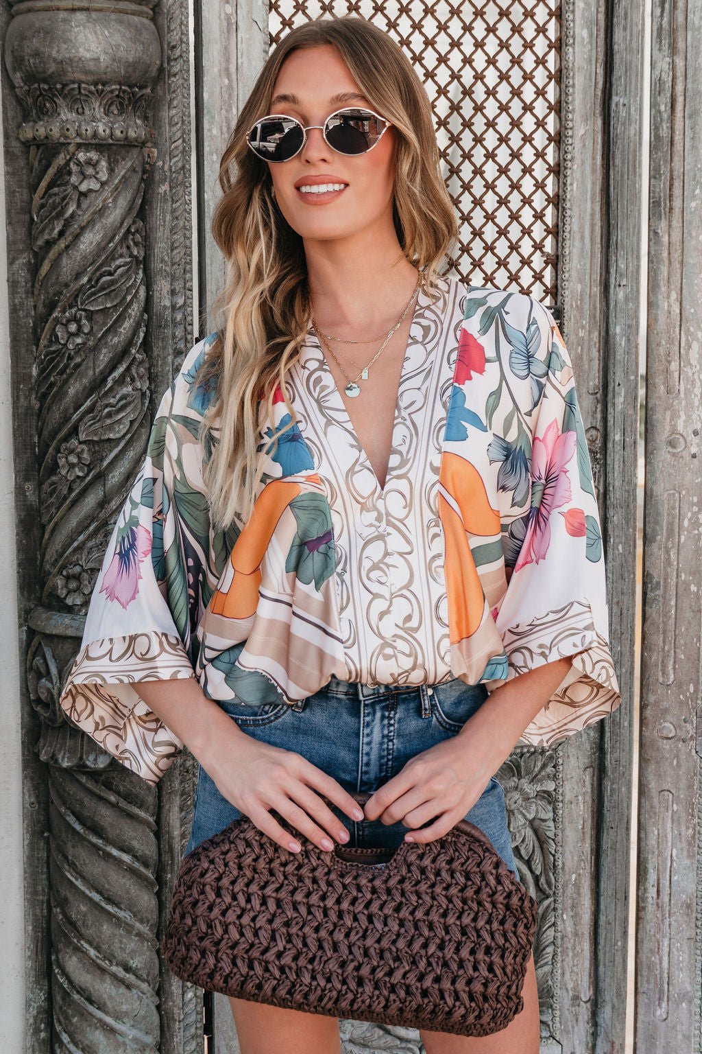 Woman in sunglasses and the Island Muse Multi Floral Bodysuit, with a denim skirt and woven clutch, stands by a decorative door.