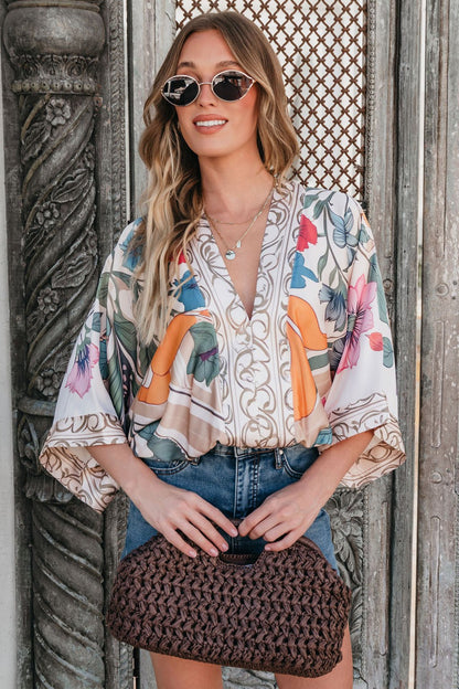 Woman in sunglasses and the Island Muse Multi Floral Bodysuit, with a denim skirt and woven clutch, stands by a decorative door.