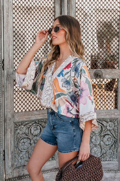 Woman in the Island Muse Multi Floral Bodysuit with kimono sleeves, denim shorts, sunglasses, and a brown woven bag by wooden doors.