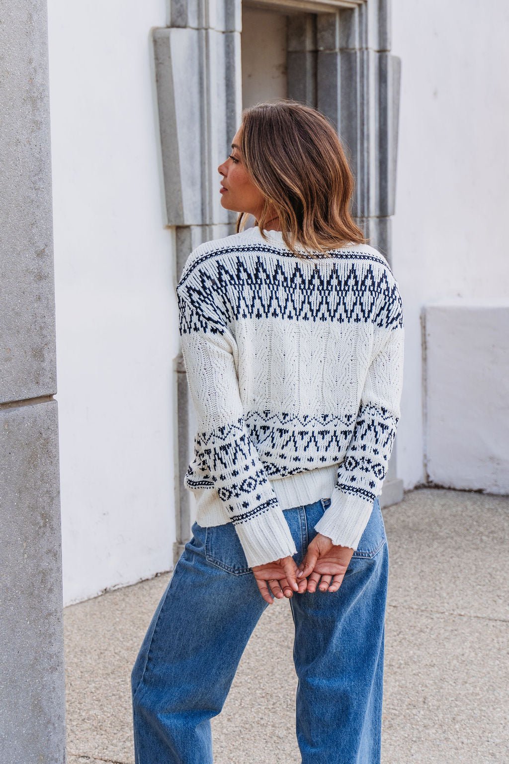 A woman wears an Ivory and Black Aztec Print Cable Knit Sweater, standing outdoors with her back to the camera.