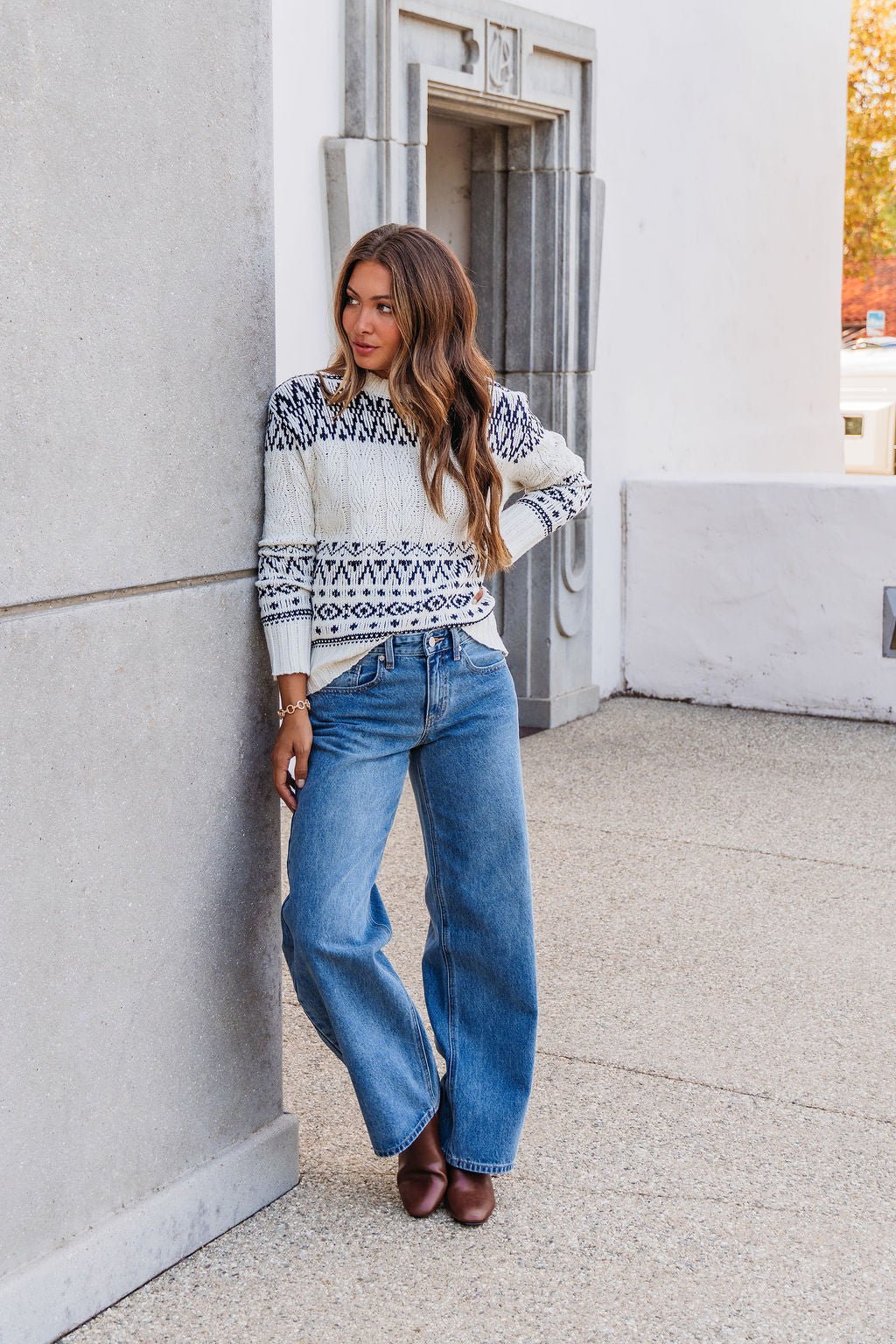 Woman with long hair leans against a wall outdoors in the Ivory and Black Aztec Print Cable Knit Sweater – FINAL SALE.