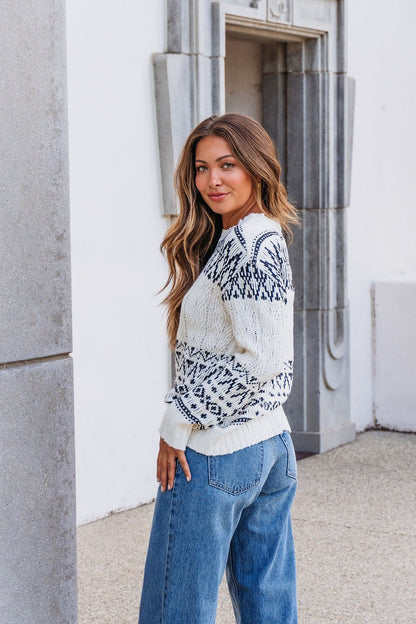 Woman with long brown hair in an ivory and black Aztec print cable knit sweater, standing outdoors near a stone building.