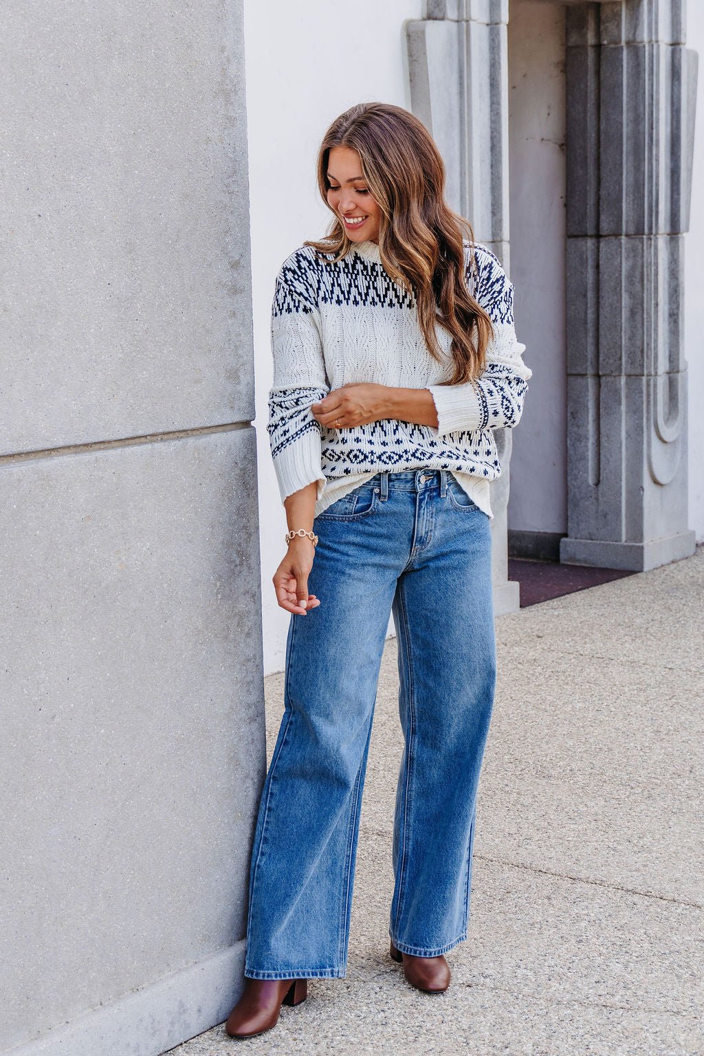 A woman in an Ivory and Black Aztec Print Cable Knit Sweater smiles near a stone wall and archway.