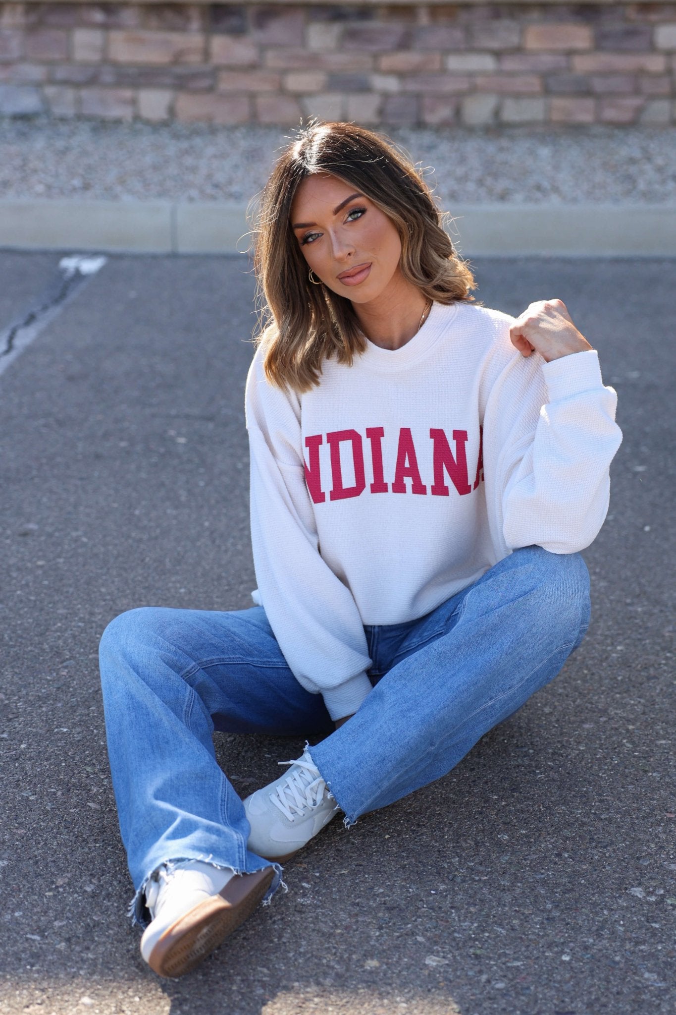 Woman sits on pavement in an ivory and red Indiana graphic pullover, blue jeans, and sneakers with a brick wall behind her.