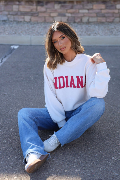 Woman sits on pavement in an ivory and red Indiana graphic pullover, blue jeans, and sneakers with a brick wall behind her.