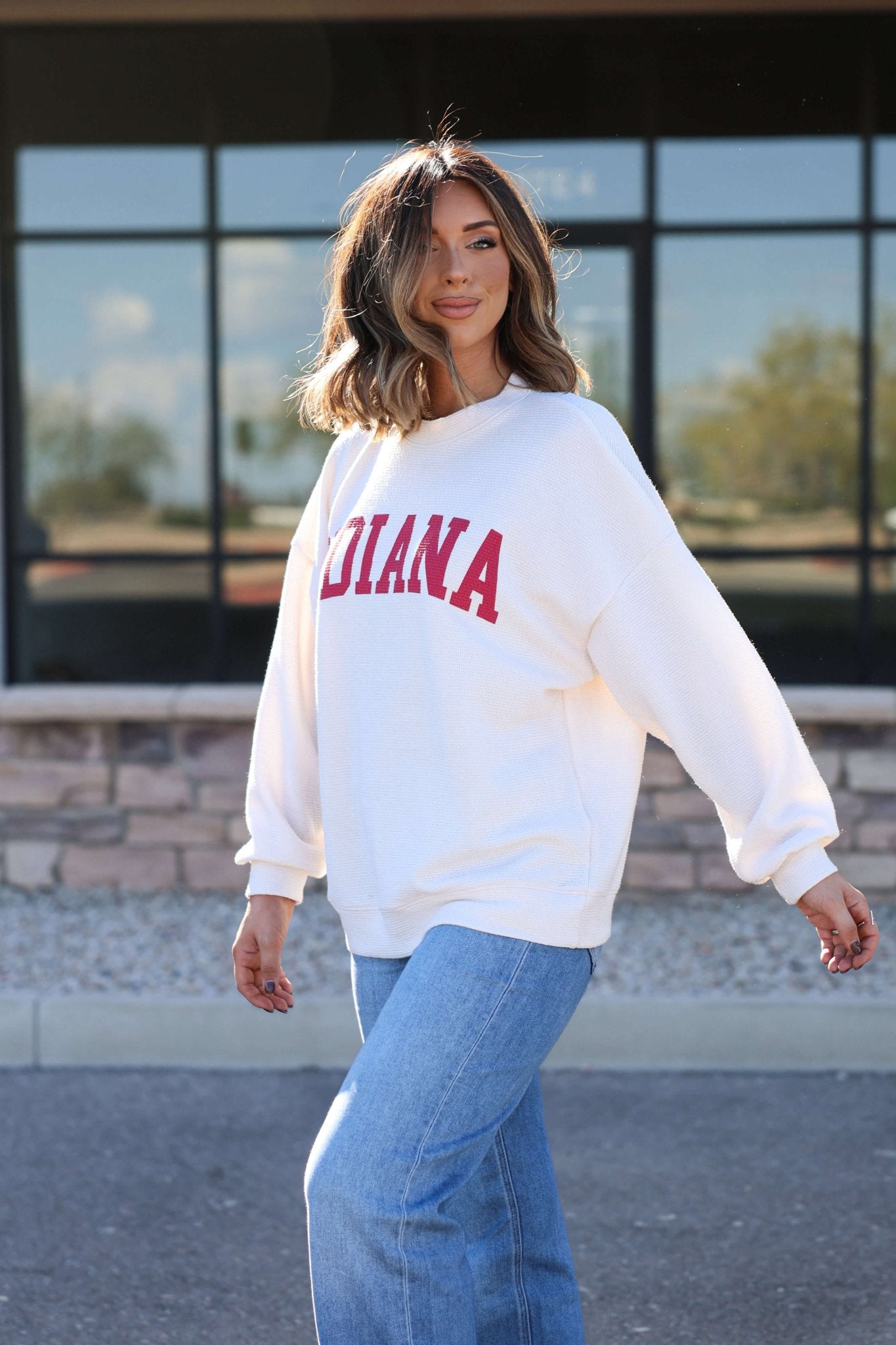 Woman with shoulder-length brown hair, smiling outdoors in an Ivory and Red Indiana Graphic Pullover and blue jeans.
