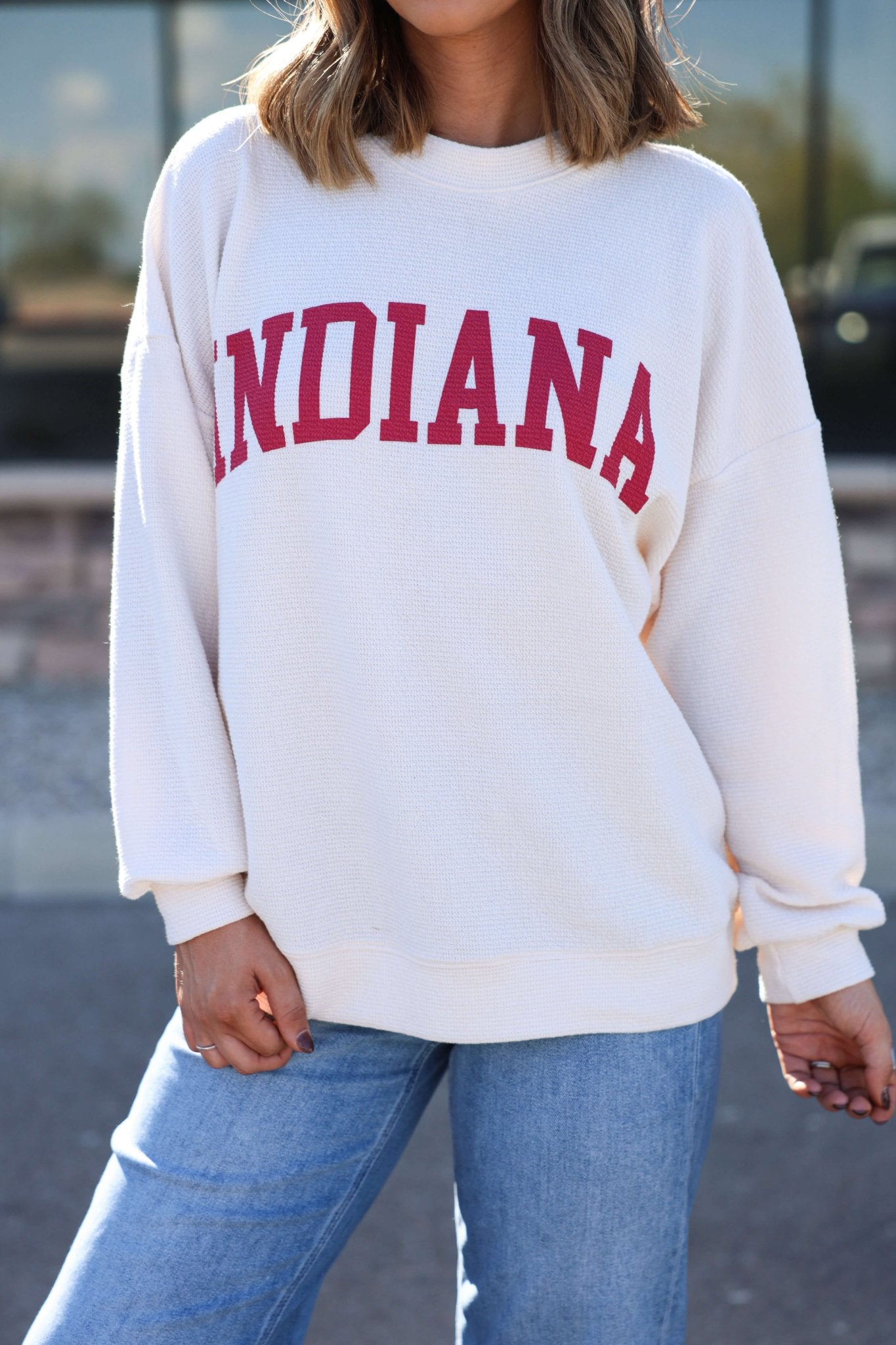 Wearing an Ivory and Red Indiana Graphic Pullover with red INDIANA letters and light blue jeans, standing outdoors.
