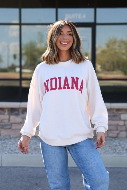 Woman smiles outdoors in an ivory and red Indiana Graphic Pullover and blue jeans, with a building in the background.