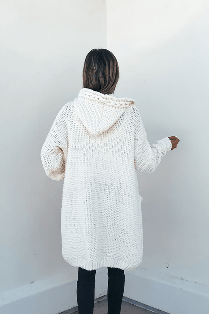 Person faces away from the camera in an Ivory Braided Trim Hooded Cardigan and black pants against a white wall.