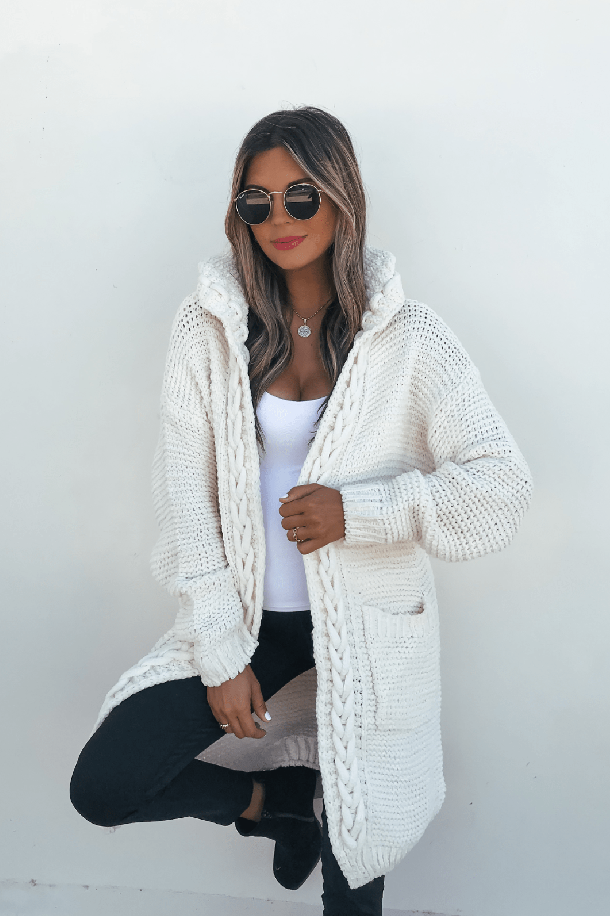 Woman wearing an Ivory Braided Trim Hooded Cardigan, black pants, sunglasses, and red lipstick against a white wall.