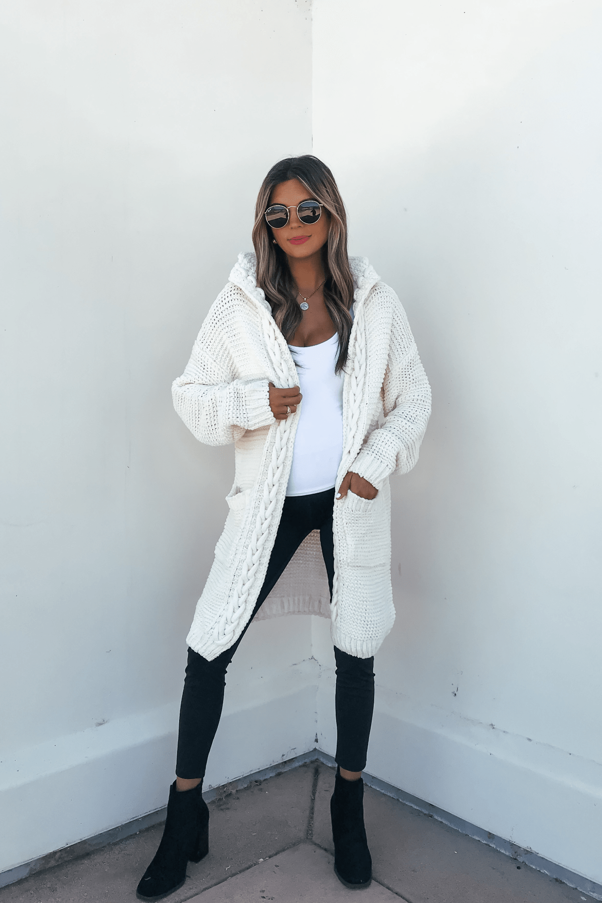 A woman in sunglasses models the Ivory Braided Trim Hooded Cardigan over a white top, black pants, and black boots.