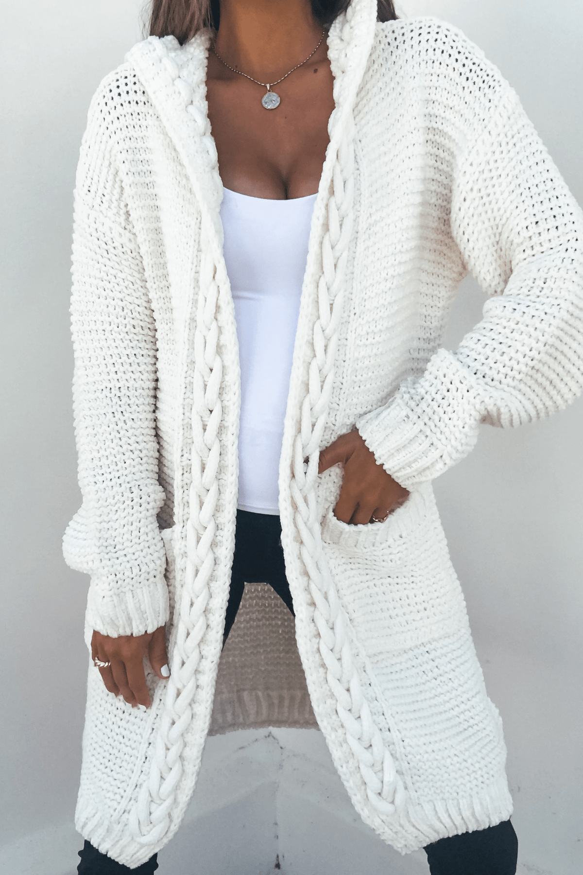 A woman wears the Ivory Braided Trim Hooded Cardigan over a white top and black pants.