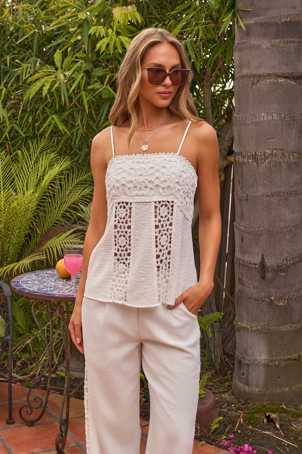 Woman wearing the Ivory Coast Crochet Top with matching white pants, sunglasses, and standing outdoors by plants and a small table.