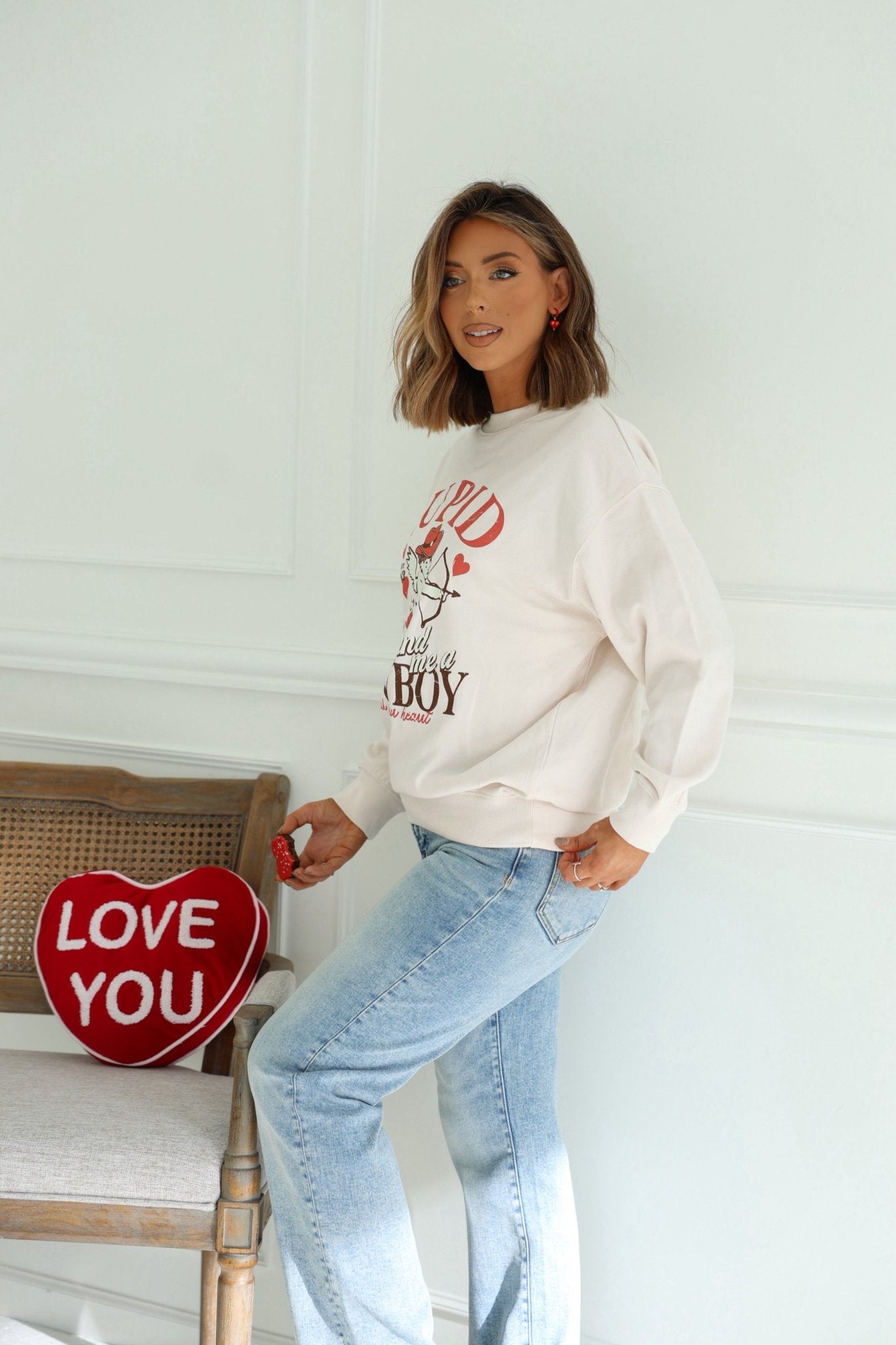 A woman in a Light Beige Cupid Cowboy Graphic Sweatshirt and blue jeans stands by a chair with a red heart-shaped LOVE YOU pillow.