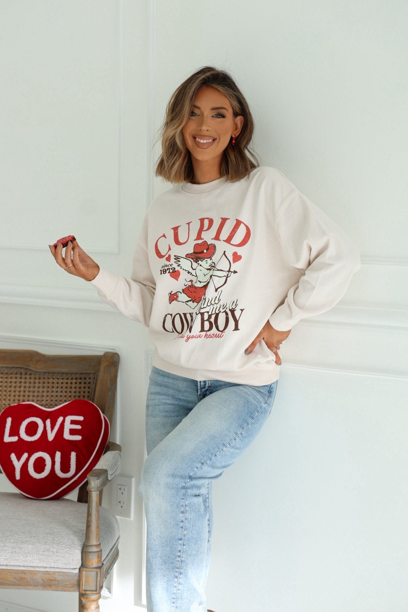 Woman smiles in a Light Beige Cupid Cowboy Graphic Sweatshirt, leaning on a chair; 100% cotton comfort.